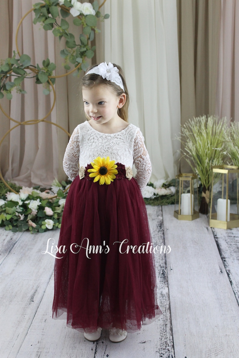 Burgundy long sleeve dress for a flower girl. A young girl wearing a white lace long-sleeved dress with a burgundy tulle skirt. The dress has a sunflower and brown flower embellishment at the waist. The girl is wearing a white headband with a white flower on top.