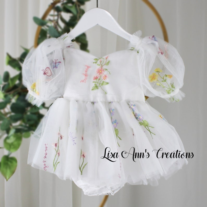 A white tulle baby dress with embroidered flowers and butterflies. The dress has a ruffled skirt and puff sleeves.