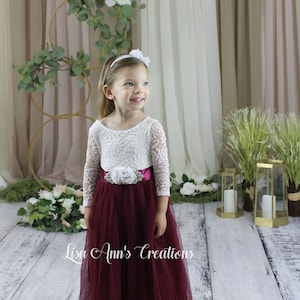 Toddler Flower Girl in a burgundy and white dress. A young girl wearing a white lace long-sleeved dress with a burgundy tulle skirt and a white flower sash. The girl is wearing a white headband with a white flower on top.