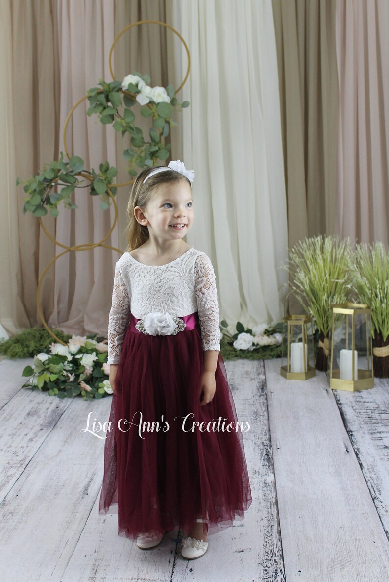 Toddler Flower Girl in a burgundy and white dress. A young girl wearing a white lace long-sleeved dress with a burgundy tulle skirt and a white flower sash. The girl is wearing a white headband with a white flower on top.