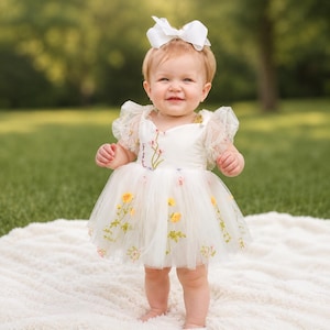May include: A white tulle dress featuring short ruffled sleeves and delicate floral embroidery in yellow, pink, and green. The dress has a full skirt and is paired with a large white hair bow, creating a coordinated ensemble for a young child.