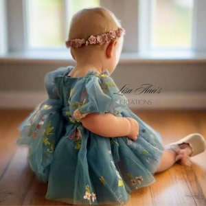 May include: A baby girl wearing a light blue tulle dress with floral embroidery and a pink flower crown. The dress has puffed sleeves and a full skirt. The baby is sitting on a wooden floor and looking to the right. The text "Lisa Ann's Creations" is visible in the background.