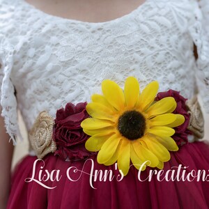 May include: A white lace dress with a maroon tulle skirt and a sunflower and burgundy flower sash. The sash is adorned with burlap fabric. The dress is labeled "Lisa Ann's Creations".