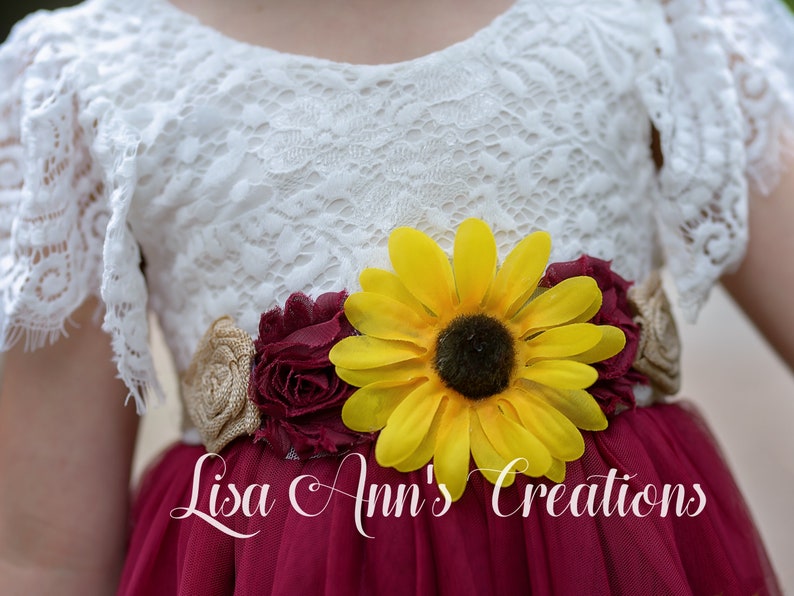 May include: A white lace dress with a maroon tulle skirt and a sunflower and burgundy flower sash. The sash is adorned with burlap fabric. The dress is labeled "Lisa Ann's Creations".