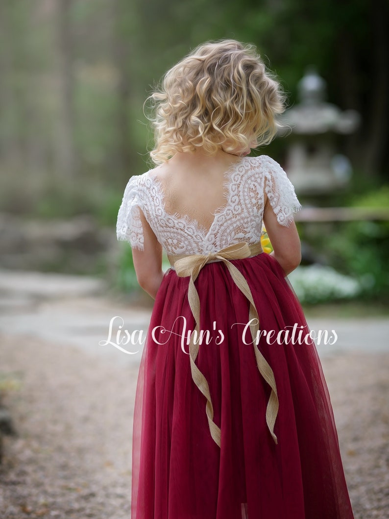 May include: A young girl wearing a white lace top with a burgundy tulle skirt and a brown sash. The skirt is long and flowing, and the sash is tied in a bow at the back. The girl is standing in a garden setting.