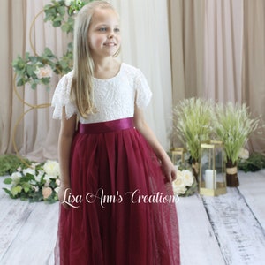 May include: A young girl wearing a white lace dress with a burgundy tulle skirt and a burgundy satin sash. The dress has short sleeves and a round neckline. The girl is standing in front of a white backdrop with greenery and flowers.