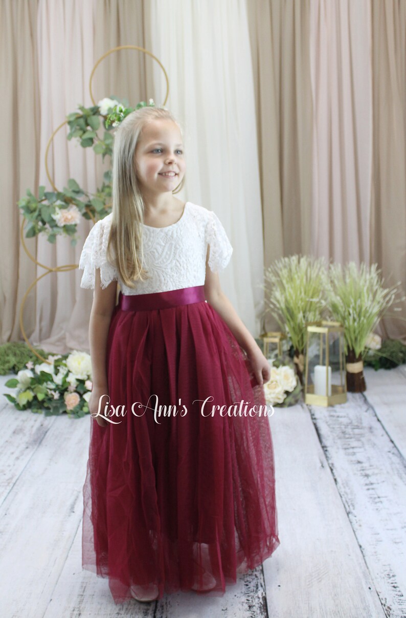 May include: A young girl wearing a white lace dress with a burgundy tulle skirt and a burgundy satin sash. The dress has short sleeves and a round neckline. The girl is standing in front of a white backdrop with greenery and flowers.