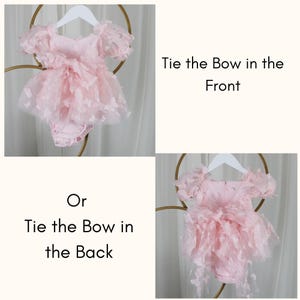 May include: A light pink romper featuring short puff sleeves and a layered tulle skirt adorned with small butterfly appliqu&eacute;s. The garment includes a long fabric sash that can be tied into a bow in the front or the back, as indicated by the text.