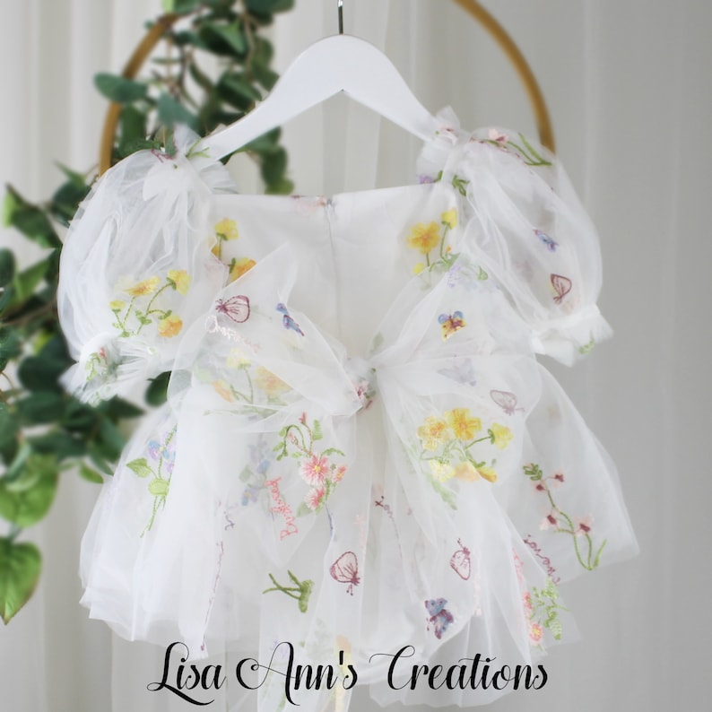 A white tulle dress with embroidered flowers and butterflies. The dress has a bow in the back and puffed sleeves.