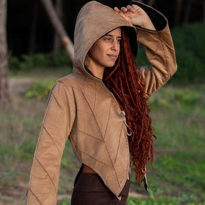 Puede incluir: Una chaqueta marrón con capucha, manga larga y caída y un dobladillo puntiagudo. La chaqueta se lleva sobre unos leggings marrones con un diseño geométrico de recortes.