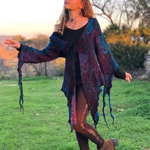 Op de afbeelding: Een vrouw draagt een lange, vloeiende, paarse en blauwe gebloemde cardigan met een zwart topje en een bruine legging. De cardigan heeft een strik in de taille en de legging heeft kant-achtige details. Ze draagt bruine laarzen en heeft lang, krullend haar.