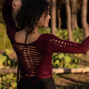Peut inclure: Une femme portant une chemise à manches longues de couleur bordeaux avec un laçage dans le dos. La chemise est faite d'un tissu doux et extensible et a une coupe décontractée. La femme est debout dans un environnement forestier.