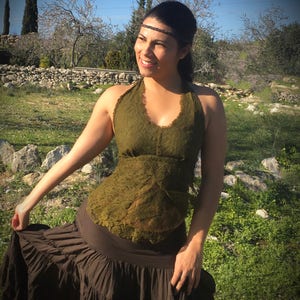 Op de afbeelding: Olijfgroene haltertop met een kanten design en een bruine rok met ruches. De vrouw draagt een zwarte haarband. De achtergrond toont een stenen muur, bomen en een grasveld.