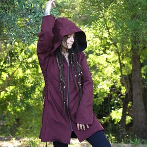 Puede incluir: Un abrigo con capucha burdeos con interior negro, cremallera frontal y lazos laterales. El abrigo se lleva con leggings y botas negras. El fondo es un exuberante bosque verde con luz solar.
