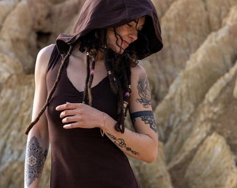 May include: A woman wearing a brown hooded dress with a long, dark braid. She has tattoos on her arms and is standing in a rocky, desert-like landscape.
