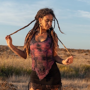 Può includere: Una donna con lunghi dreadlocks marroni indossa un top corto tie-dye con un bordo in pizzo nero e una minigonna nera con un bordo in pizzo. È in piedi in un campo di erba secca.