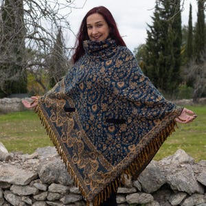 Op de afbeelding: Een blauwe en gouden poncho met franjes. De poncho heeft een hoge hals en twee zakken.