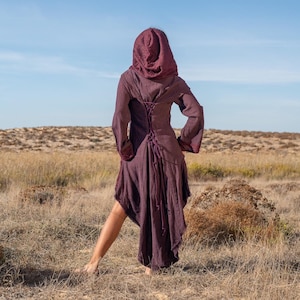 May include: A woman wearing a long, hooded, burgundy dress with a lace-up back. She is standing in a field of dry grass with a blue sky in the background.