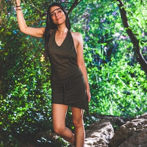 May include: A woman wearing a dark green halter dress stands in a woodland setting. She is smiling and looking at the camera. The dress has a drawstring waist and a flowing skirt.
