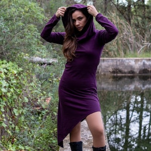 Könnte beinhalten: Eine Frau trägt ein langes, violettes Kapuzenkleid mit hohem Kragen und einem langen, fließenden Rock. Sie steht auf einem Betonweg in der Nähe eines Gewässers.
