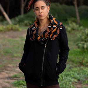 Puede incluir: Una mujer con rastas rojas lleva una chaqueta negra con cremallera y una bufanda con estampado marrón y negro. La chaqueta tiene una cremallera y un cuello.