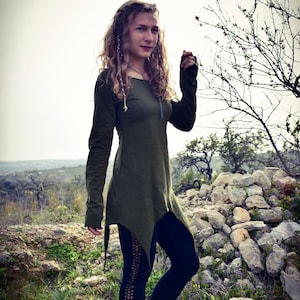 May include: A woman wearing a long-sleeved, green, asymmetrical tunic top with a scoop neck. The top has a flowing, layered design and is made of a soft, comfortable fabric. The woman is standing in a natural setting with rocks and trees in the background.