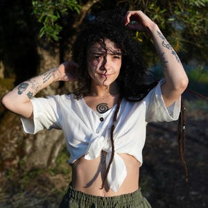 Op de afbeelding: Een witte top met korte mouwen en een V-hals, met twee zwarte knopen. De top is geknoopt. De persoon draagt een olijfgroene broek. De persoon heeft donker krullend haar en dreadlocks. Tatoeages zijn zichtbaar op de armen en borst.