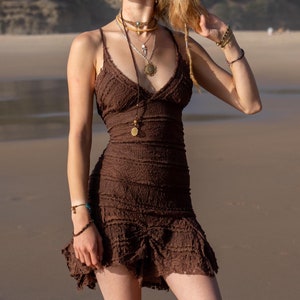 Peut inclure: Une femme aux longs cheveux blonds portant une robe en crochet marron et plusieurs colliers en or avec des pendentifs. Elle se tient sur une plage de sable avec une colline verte en arrière-plan.