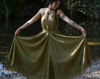 May include: Olive green halter dress with a flowing skirt and geometric pattern near the hem. The dress has a cinched waist and is worn outdoors near a body of water.