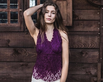 May include: A woman wearing a purple lace halter top and a white skirt stands in front of a wooden wall. The top has a V-neck and a scalloped edge. The skirt is long and flowing. The background is a rustic wooden structure.