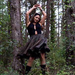 May include: A black bodysuit with a keyhole cutout paired with a brown and gray skirt with a black fringe hem. The skirt is flared, creating movement. The outfit is worn outdoors in a forest setting.