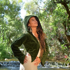 Pode incluir: Uma jaqueta verde de veludo cortada com capuz e mangas compridas. A jaqueta tem um design acolchoado e está sendo usada por uma pessoa com longos dreadlocks.