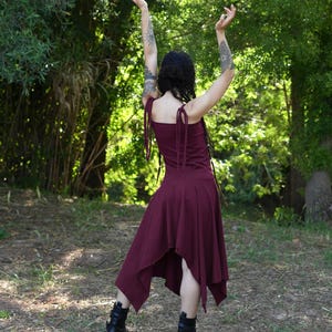 May include: A burgundy dress with a flowing skirt and tie straps. The dress is paired with black boots. The person is standing in a forest with arms raised.