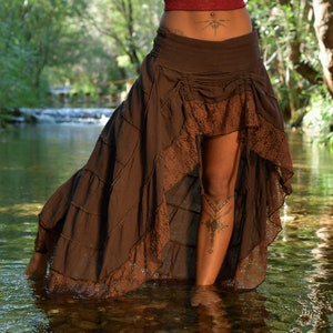 May include: A brown, tiered skirt with lace detailing. The skirt is asymmetrical, with a longer back and a shorter front. The skirt is worn by a person standing in a shallow stream.