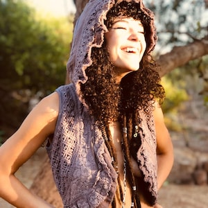 May include: A woman with curly brown hair is wearing a purple crochet hooded vest with a ruffled neckline. She is smiling and looking up.