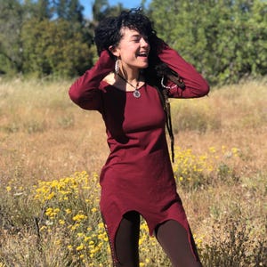 Peut inclure: Une tunique bordeaux à manches longues avec un ourlet asymétrique unique. La tunique est associée à des leggings marron foncé. La tenue est portée à l'extérieur dans un champ de fleurs sauvages jaunes et d'herbes hautes.