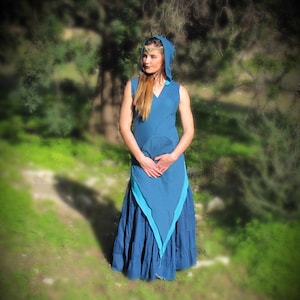 May include: A sleeveless, blue hooded dress with a long, flowing skirt. The dress features a V-neck and a contrasting turquoise panel. A gold-colored headpiece adorns the head. The person is standing in a grassy area with trees in the background.
