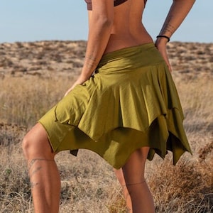 Puede incluir: Una mujer con rastas largas lleva un top marrón de halter y una falda verde de capas. Está de pie en un campo de hierba alta con un cielo azul de fondo.