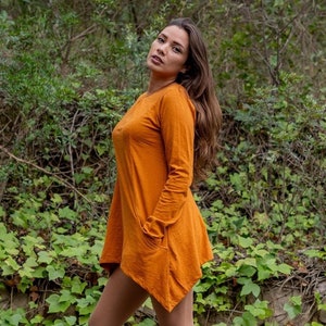 May include: A woman wearing a long-sleeved, rust-coloured, asymmetrical dress with a pocket on the left side. She is wearing white lace-up boots and standing in front of a wall covered in green vines.