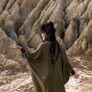 May include: A woman with long, dark hair wearing a long, olive green, loose-fitting cardigan with a textured weave. She is standing in a desert landscape with a rocky, eroded background.