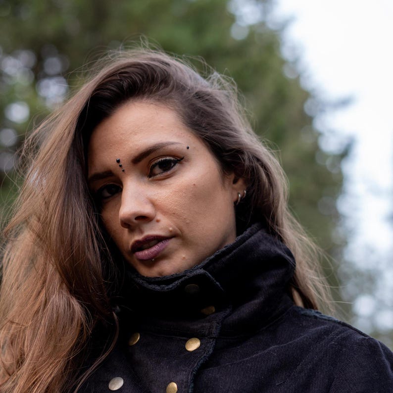 May include: A woman with long brown hair wearing a black jacket with gold buttons. She has a small black dot above her eyebrow.