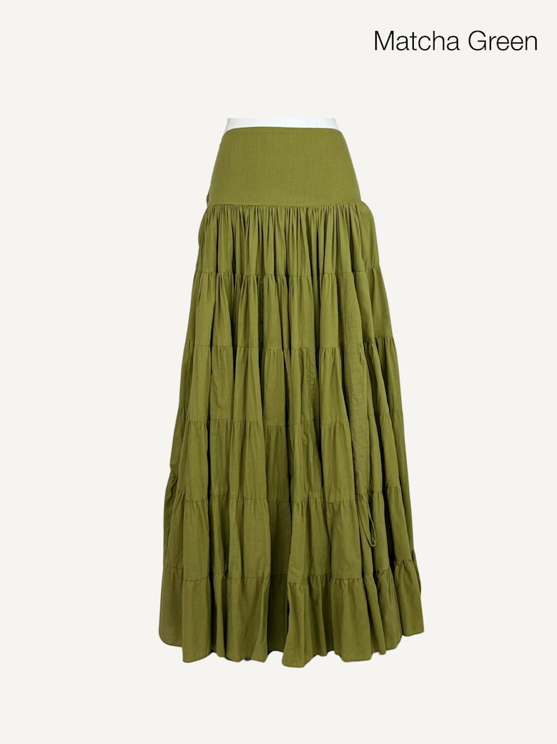 May include: Matcha green tiered maxi skirt. The skirt features a high waist and multiple layers of fabric, creating a flowing silhouette. The skirt is a solid color and has a drawstring closure. The text "Matcha Green" is in the upper right corner.