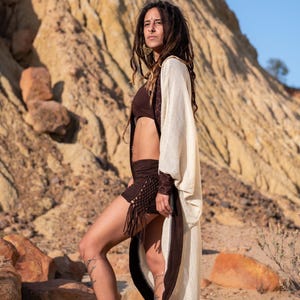 May include: A woman wearing a brown crop top, shorts with fringe, and a cream-colored kimono with brown trim. The kimono has a geometric pattern on the sleeves. The background is a desert landscape with rock formations.