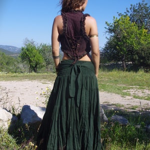 May include: A dark green tiered maxi skirt paired with a burgundy lace-up vest. The skirt has a drawstring waist and a flowing silhouette. The vest features a ruffled trim and a laced back. The outfit is worn outdoors in a natural setting.