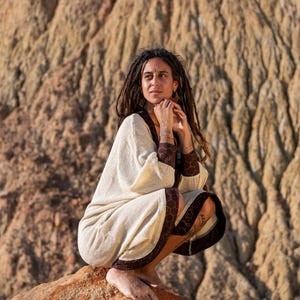 May include: A cream-colored robe with brown hexagonal trim and cuffs. The robe has wide sleeves and a relaxed fit. The model is wearing the robe while sitting on a rock. The background is a blurred, textured rock formation.