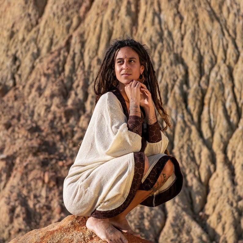 May include: A cream-colored robe with brown hexagonal trim and cuffs. The robe has wide sleeves and a relaxed fit. The model is wearing the robe while sitting on a rock. The background is a blurred, textured rock formation.