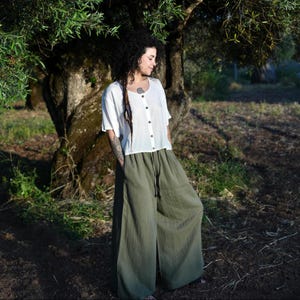 Puede incluir: Una camisa blanca abotonada de manga corta y pantalones de pierna ancha verde oliva. Los pantalones tienen cintura con cordón. El conjunto está hecho de una tela ligera, perfecto para el clima cálido. El modelo está delante de un gran árbol.
