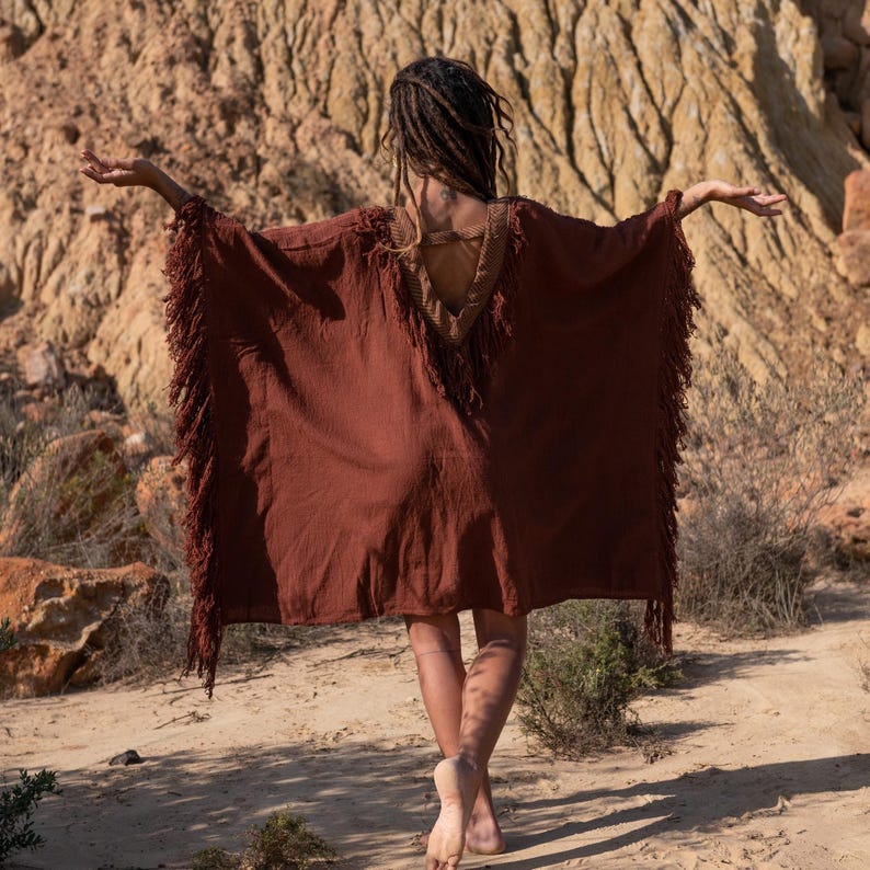 May include: A rust-colored poncho with fringed edges and a decorative neckline. The open-back design and flowing fabric create a bohemian aesthetic. The model is standing outdoors with arms outstretched.
