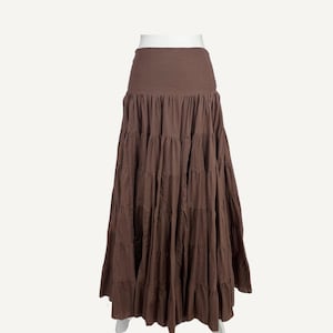 May include: Mauve tiered maxi skirt. The skirt features a wide, solid-colored waistband and multiple horizontal tiers that create a flowing, full silhouette. The skirt is a solid mauve color and is displayed on a white background.