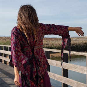 Puede incluir: Una mujer lleva una blusa de manga larga con estampado floral morado y azul con lazada en la espalda. La blusa tiene un dobladillo asimétrico y las mangas tienen bordes con volantes.